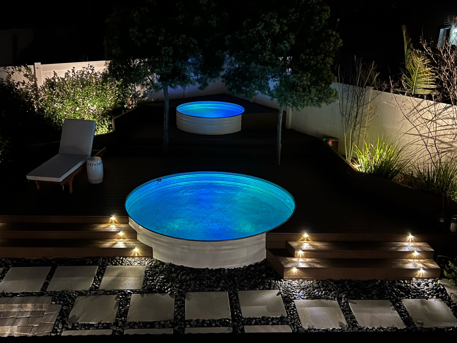 Two round stock tank pools in a garden setting at night with illuminated pathways.
